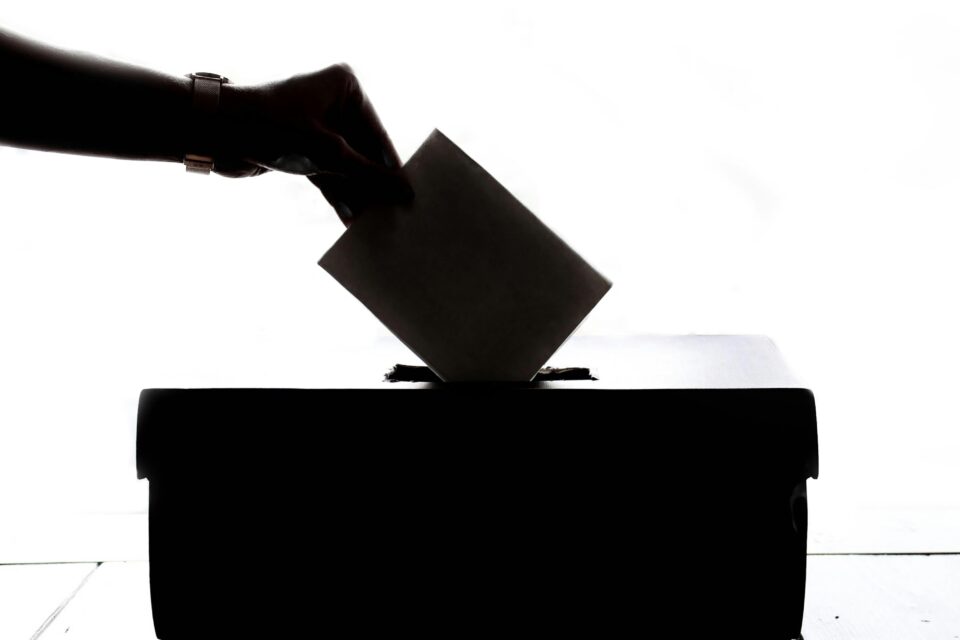 In nero su fondo bianco, una mano inserisce una scheda in un'urna elettorale.